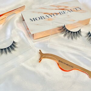 Op de afbeelding: Twee paar valse wimpers in doorzichtige plastic doosjes, een wimperapplicator van rosé goud en een rosé gouden doos met de tekst "MORAYO BEAUTY" op de deksel.