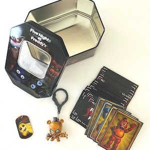 May include: An open, black, octagonal tin featuring the text "Five Nights at Freddy's" and character images. Includes a keychain, dog tag, and a stack of cards with character illustrations. The tin is empty.
