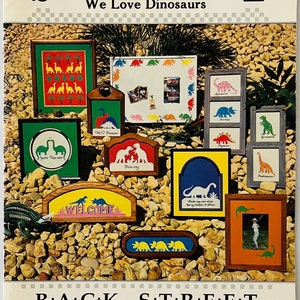 May include: A book titled "Scherenschnitte We Love Dinosaurs" with a cover featuring various wooden signs with dinosaur designs. The signs are decorated with colourful paint and feature different dinosaur silhouettes, including a Tyrannosaurus Rex, a Stegosaurus, and a Triceratops. The signs are arranged on a background of small, brown stones.