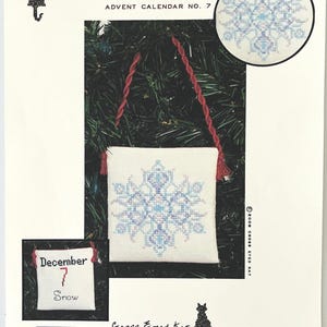 May include: A cross-stitch Advent calendar kit featuring a snowflake design. The kit includes a small, white fabric pouch with a blue and white snowflake pattern, red braided cord, and a small calendar with the text "December 7 Snow".