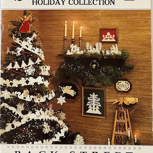 May include: A book cover titled "SCHERENSCHNITTE HOLIDAY COLLECTION" featuring a decorated Christmas tree with paper cutouts, framed art, and a wooden pyramid. The cover also displays the text "BACK STREET DESIGNS".