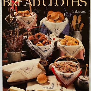 May include: A vintage needlepoint leaflet featuring 9 bread cloth designs. The cover shows a variety of breads in baskets and bowls, with embroidered cloths featuring sayings like "Give us this day our daily bread" and "Home sweet home".