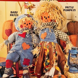 May include: A McCall's Creates sewing pattern for two pumpkin bumpkin dolls. The pattern is number 15087. The dolls are dressed in denim overalls and a plaid skirt with autumn-themed fabric. The dolls are sitting on bales of straw with a lantern and a toy truck.