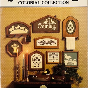 May include: A collection of wooden signs with various designs, including a pineapple, a house, a duck crossing, and the words "Country" and "Home Sweet Home". The signs are displayed on a brown surface with candles and a wooden duck decoy.