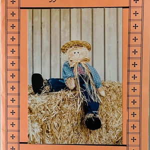 Puede incluir: Un patrón para una muñeca de espantapájaros de 50 cm, llamada "Johnny Hayseed", de la línea Stuff-n-Stitches. El patrón presenta una foto a color de una muñeca de espantapájaros terminada sentada en una paca de paja.