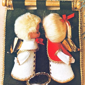 Puede incluir: Un adorno navideño con dos muñecas ángeles. Tienen cabello rubio, vestidos blancos y alas doradas. Un ángel lleva un chaleco rojo. El adorno está enmarcado con adornos dorados y sobre un fondo verde oscuro.