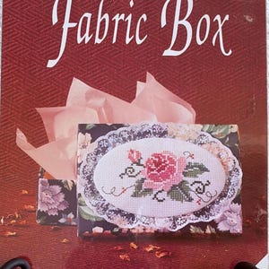 May include: A white ceramic mug with a black handle and the text "But First, Coffee" in a bold, black font. The mug is set against a blurred background. The mug appears to be approximately 10 cm tall.