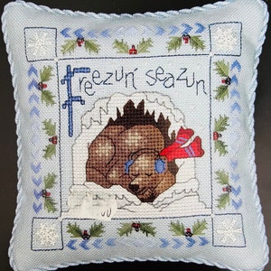 Może przedstawiać: Poduszka haftowana w kolorze niebieskim i białym z brązowym niedźwiedziem w czerwonej czapce i niebieskich nausznikach. Poduszka ma białą ramkę i wzór płatka śniegu w rogach. Tekst "Freezin' season" jest wyszyty w środku.