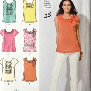 Peut inclure: Patron de couture Simplicity 2599 présentant divers modèles de hauts avec des volants et des nœuds. Le patron comprend les tailles 4, 6, 8, 10 et 12. L'image montre également une femme portant un haut orange et un pantalon blanc.