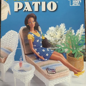 May include: A cover for a "Fashion Doll Patio" plastic canvas project. The image shows a doll in a blue and white dress, seated on a woven sun lounger. A small table and a potted plant are also visible. The text "Fashion Doll Patio" is displayed.