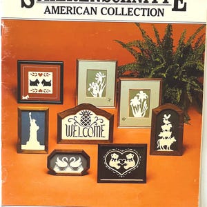 May include: The cover of a book titled "SCHERENSCHNITTE AMERICAN COLLECTION" features framed paper cut art. The art includes images of the Statue of Liberty, swans, dogs, and floral designs. The background is orange with a fern.