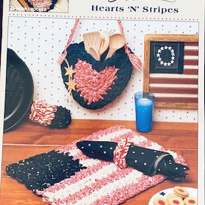 May include: A craft pattern book titled "McCall's Hearts 'N' Stripes" featuring crochet designs. The image showcases various patriotic-themed crochet items, including a heart-shaped hanging basket, a placemat, and a napkin ring, all in red, white, and blue.