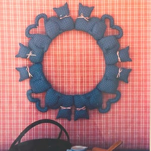 May include: A blue cat-shaped wreath with white polka dots and pink bows. The wreath is surrounded by a black basket, a small blue pitcher, a wooden spoon, and a rolling pin. The background is a pink and white plaid pattern.