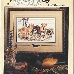 May include: A framed print titled "The Little Bandits" by Phillip Crowe, featuring a cross-stitch image of three golden retriever puppies with ducks. The image includes a decorative wooden duck decoy and a tin container.
