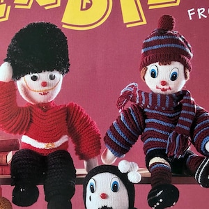 May include: Two knitted dolls are featured, one dressed as a British Royal Guard in a red jacket and black hat, the other in a striped sweater, hat, and scarf. Both dolls have large eyes, red noses, and smiling mouths. The dolls are sitting on a shelf.