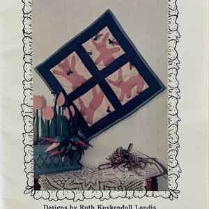 May include: A quilt pattern for a 14-inch by 14-inch quilt featuring four pink bunnies with blue accents. The quilt is titled "Juggling Bunnies" and is designed by Ruth Kuykendall Landis.