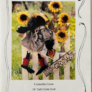 May include: A sewing pattern for an 18-inch tall cloth doll called Cornelia Crow. The doll is wearing a plaid dress and red boots. The pattern is by Designs by Madonna and is numbered DbM 46.