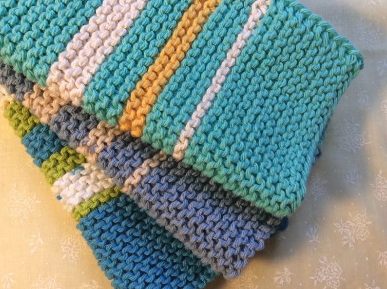 May include: A stack of four knitted dishcloths in various shades of blue, green, and white. The dishcloths are made with a textured stitch and have a striped pattern.
