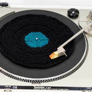 May include: A black and teal knitted record sits on a silver turntable with a tone arm and needle. The turntable is labelled "Technics SL-BD20 Frequency Generator Servo Automatic Turntable System".