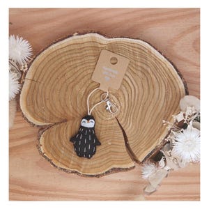 Könnte beinhalten: Ein handgefertigter Pinguin-Schlüsselanhänger mit schwarzem Körper und weißem Bauch, mit einem kleinen Anhänger mit der Aufschrift "Handmade with love". Der Schlüsselanhänger ist an einem silbernen Verschluss und einer Kordel befestigt, auf einer Holzscheibe.