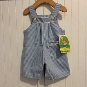 May include: Blue and white striped baby overalls with white trim and button details. The overalls are hanging on a wooden hanger. A tag with the text "Mufflings" is attached to the overalls.