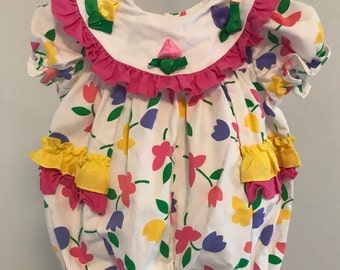 May include: White baby romper with a floral pattern of pink, yellow, and purple flowers. The romper has a pink ruffled collar and trim, with yellow and pink ruffled accents on the front. It is hanging on a clothesline.