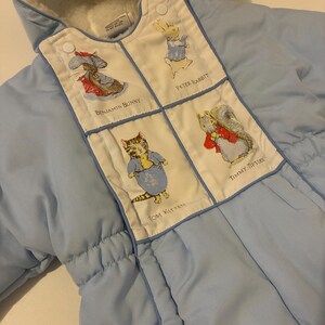 May include: A light blue hooded snowsuit with a white fleece lining. The front features a white panel with four illustrated characters: Peter Rabbit, Benjamin Bunny, Tom Kitten, and Timmy Tiptoes, each labeled with their respective names.