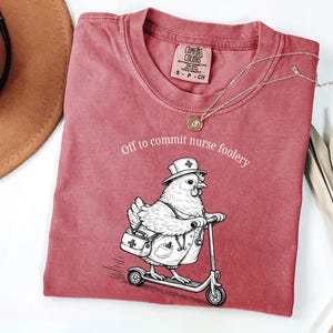 May include: A dusty rose Comfort Colors t-shirt with the phrase "Off to commit nurse foolery" and a graphic of a chicken dressed as a nurse riding a scooter. A silver necklace and a brown hat are also visible.