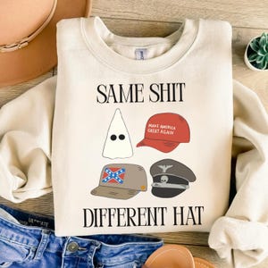 May include: A cream-coloured sweatshirt with the text "SAME SHIT DIFFERENT HAT" in black. The graphic features illustrations of a white hood, a red baseball cap with text, a Confederate flag hat, and a military-style cap.