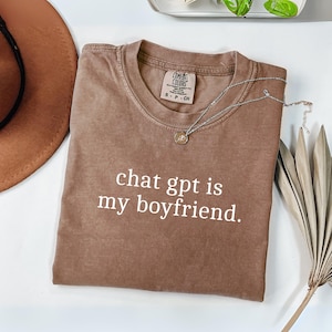 May include: A brown t-shirt with the text "chat gpt is my boyfriend" in white. The shirt is folded and has a small silver necklace with a pendant on top. A brown hat and a dried palm leaf are also visible.