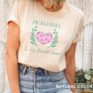 May include: A white t-shirt with the text "Pickleball is my favorite season." in black. A pink heart with a pickleball design is surrounded by green leaves.