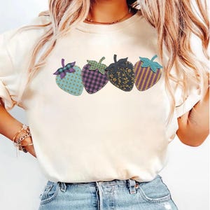 May include: A cream-coloured t-shirt with four strawberry designs. Each strawberry has a different pattern, including polka dots, plaid, floral, and stripes. The strawberries are in shades of blue, purple, and gold.