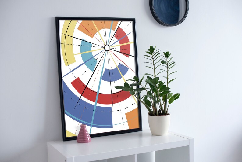 May include: A framed abstract art print with a radial design in red, blue, yellow, and orange. The artwork is displayed on a white shelf, alongside a potted plant and a small pink vase. The black frame complements the colourful design.