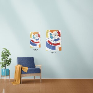 May include: Two abstract art prints with a circular design in shades of red, blue, yellow, and orange are displayed on a light blue wall. The prints are labeled 12 x 18 inches and 18 x 24 inches. A blue armchair and a potted plant are in the foreground.