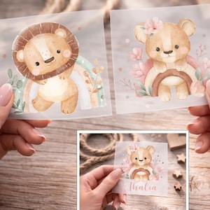 Peut inclure: Deux autocollants carrés avec des illustrations à l'aquarelle d'un lion et d'un ourson. Le lion a une crinière brune et est assis sur un arc-en-ciel. L'ourson porte une couronne de fleurs et est assis sur un arc-en-ciel. Un troisième autocollant montre l'ourson avec le nom "Thalia".