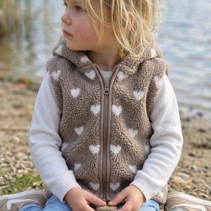 Könnte beinhalten: Eine beige Kapuzenweste aus strukturiertem Fleece-Material mit einem durchgehenden Muster aus kleinen weißen Herzen. Die Weste hat einen mittigen Reißverschluss und wird über einem schlichten weißen Langarmshirt sowie einer hellblauen Jeans getragen.