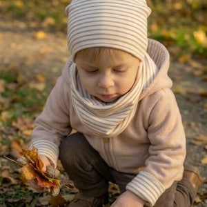 Puede incluir: Un niño con una chaqueta con capucha rosa claro, gorro y bufanda a rayas y pantalones marrones, jugando con hojas de otoño caídas. El gorro y la bufanda tienen un estampado de rayas beige y blanco. El niño está en cuclillas, rodeado de hojas coloridas.