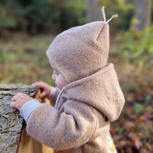 Könnte beinhalten: Eine hellbraune, texturierte Kapuzenjacke aus Wollmischgewebe mit einer spitz zulaufenden Kapuze im Wichtelstil, die an der Spitze mit zwei kleinen, hellen Zierbändern versehen ist. Das Kleidungsstück hat lange Ärmel mit hellen Bündchen.