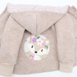 Könnte beinhalten: Eine hellbraune Kapuzenjacke mit einem Fuchs-Applikationen auf der Vorderseite. Der Fuchs trägt eine Blumenkrone und ist von einem Kreis aus Blumen umgeben.