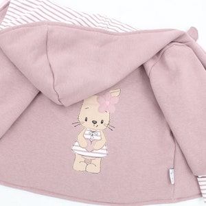 Könnte beinhalten: Mauve Kapuzenjacke mit gestreiftem Futter und Bündchen. Auf der Rückseite befindet sich ein gesticktes Cartoon-Kaninchen, das ein Kleid trägt und ein Herz hält. Die Jacke ist für Kinder konzipiert.