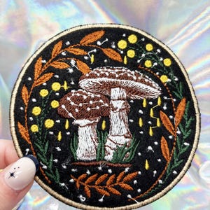 May include: Embroidered patch featuring a pair of brown and white mushrooms with white spots, surrounded by green and orange leaves and yellow flowers on a black background.