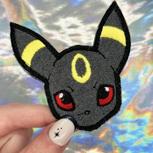 May include: Embroidered patch featuring a stylized Umbreon character with black, yellow, and red details. The patch has a detailed, stitched design with a black border and is held by a hand with a manicure.