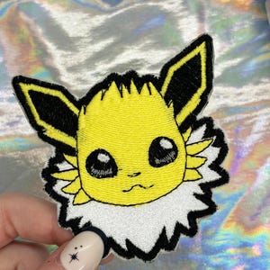 May include: Yellow and black embroidered iron-on patch of a cartoon character with large eyes and a fluffy white collar.