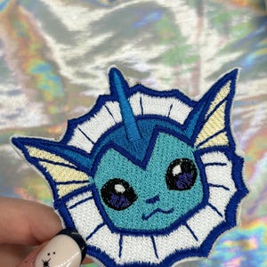 May include: A blue and white embroidered iron-on patch featuring a cartoon character with large eyes and a fin. The character is surrounded by a white scalloped border.