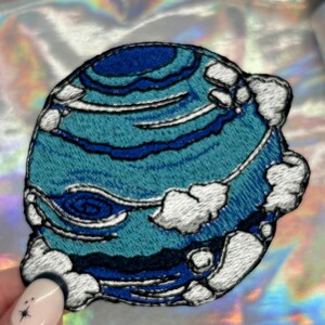 May include: A blue and white embroidered patch of a planet with clouds. The planet has a swirling pattern and is surrounded by white clouds.