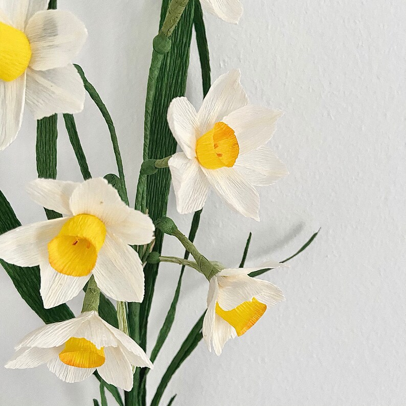 Peut inclure: Gros plan sur des jonquilles en papier faites &agrave; la main. Les fleurs ont des p&eacute;tales blancs et des centres jaune vif. Des tiges et des feuilles vertes sont visibles, dispos&eacute;es sur un fond blanc. Les fleurs en papier sont un article artisanal.