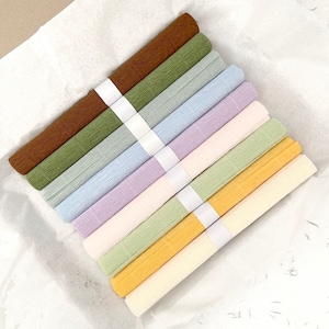 May include: A collection of rolled crepe paper in various pastel colors, including brown, green, blue, lavender, cream, and yellow. Each roll is secured with a white paper band. The paper is arranged on a white, textured surface.
