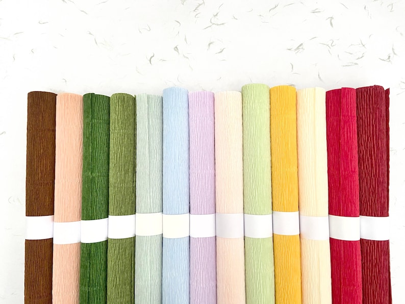 May include: A collection of rolled crepe paper in various colors, including brown, peach, green, blue, lavender, yellow, pink, and red. Each roll is secured with a white band. The paper has a textured surface, ideal for crafts and decorations.