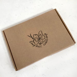 Peut inclure: Bo&icirc;te rectangulaire en carton brun avec un logo floral portant l'inscription "Campbell Workshop" dans un design circulaire. La bo&icirc;te est ferm&eacute;e et pos&eacute;e sur une surface blanche textur&eacute;e. Le logo est imprim&eacute; &agrave; l'encre noire.
