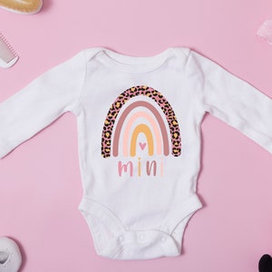 K&ouml;nnte beinhalten: Wei&szlig;er lang&auml;rmliger Baby-Strampler mit einem rosa, orangefarbenen und gelben Regenbogen mit Leopardenmuster. Der Regenbogen hat ein kleines pinkfarbenes Herz in der Mitte. Das Wort "mini" ist unter dem Regenbogen gedruckt.